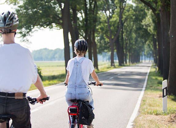 Zwei Personen mit Fahrradhelmen fahren auf einer schmalen Landstraße ohne separaten Radweg.